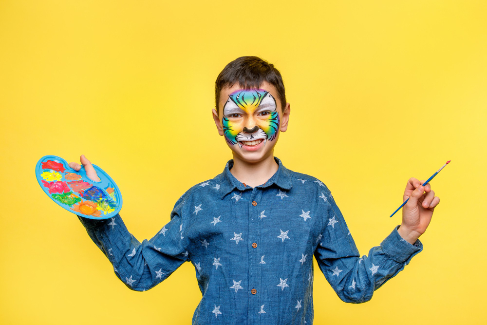 Happy boy with paint on birthday party, colorful tiger holding palette with gouache isolated on yellow wall.